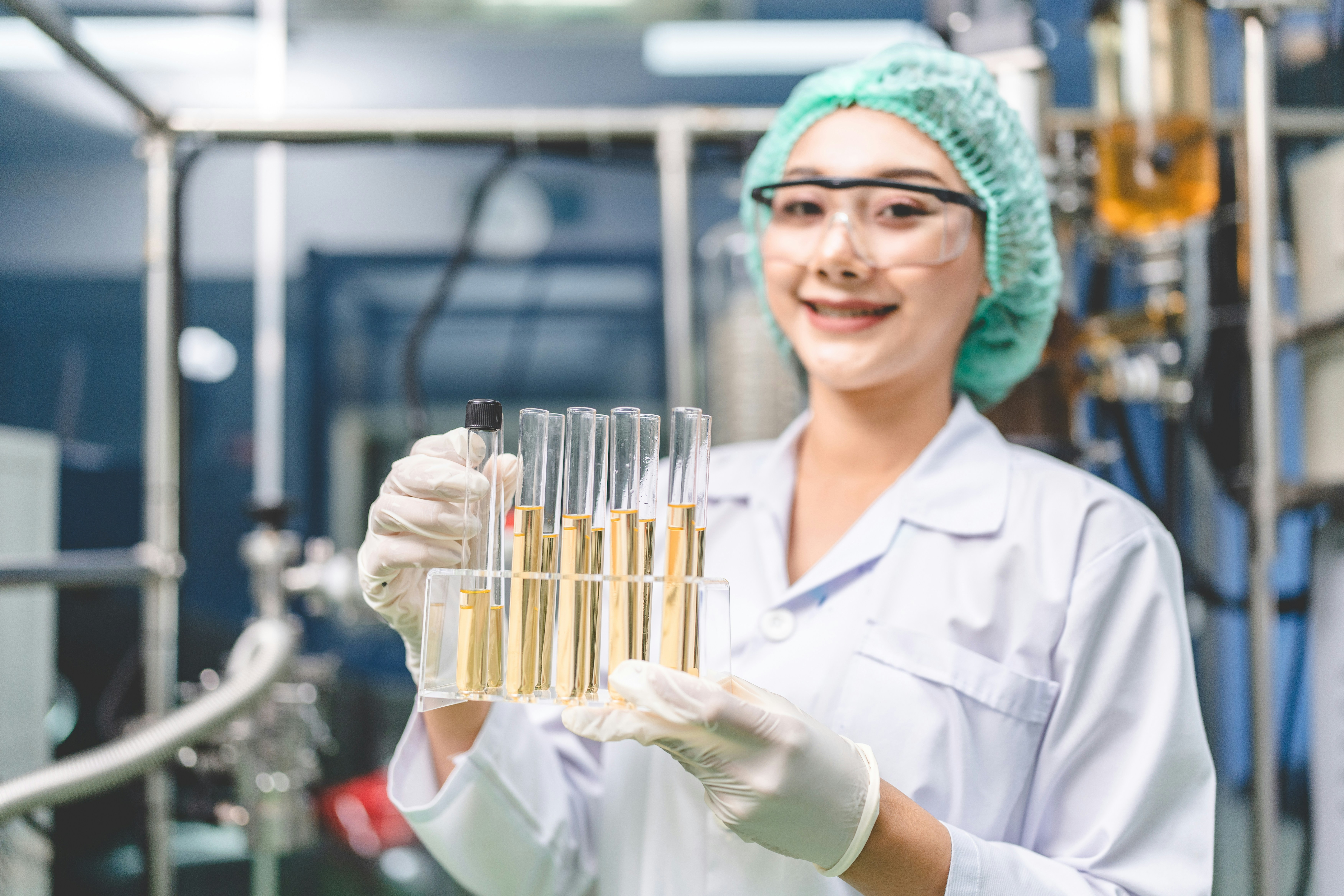 Lächelnde Laborantin hält Reag enzgläser mit gelber Flüssigkeit und trägt Laborkittel und Schutzbrille